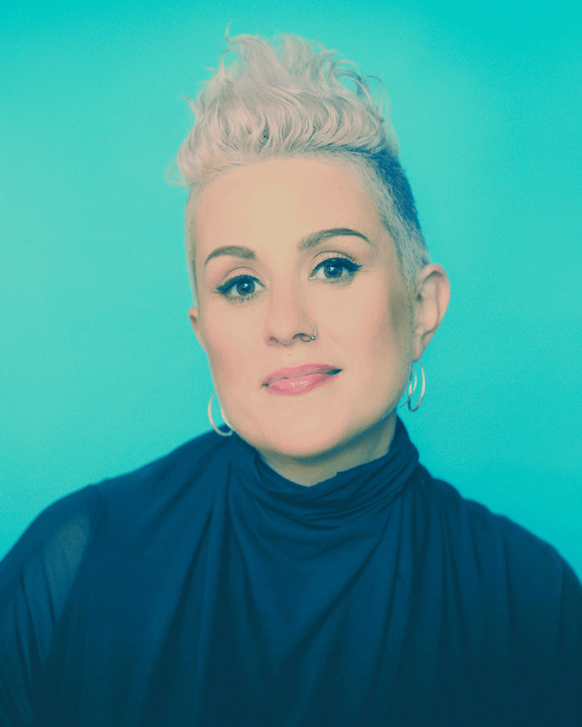 A woman with short, platinum blonde hair styled upwards, wearing hoop earrings and a dark high-neck top, poses against a bright turquoise background.