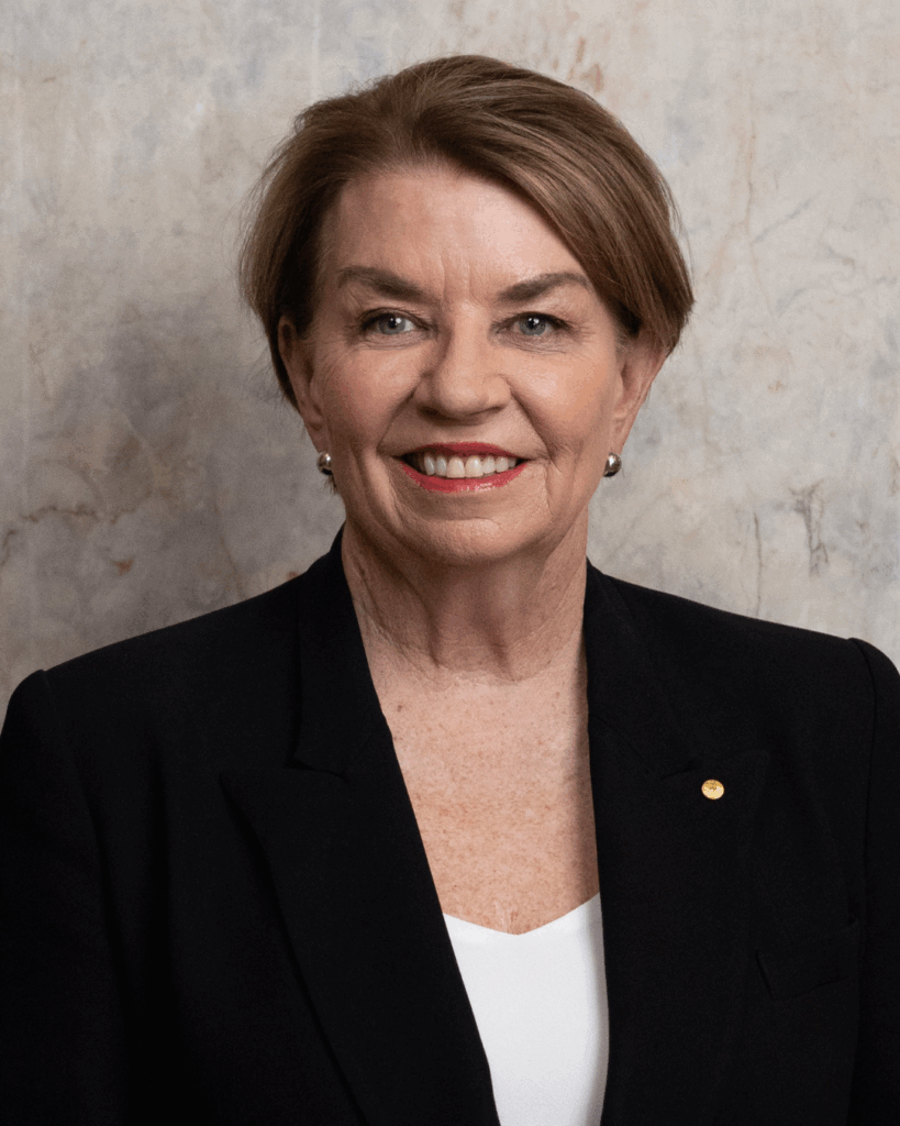 A woman with short brown hair in a black blazer, white top, and pearl earrings smiles against a marble-patterned background.