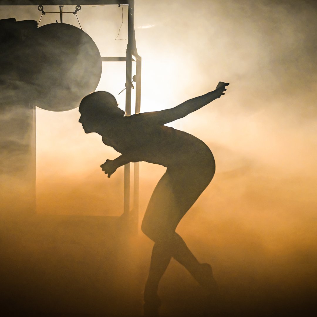 A person in a bodysuit is dramatically posing in silhouette against a bright, foggy background with industrial-looking objects and scaffolding faintly visible around them.