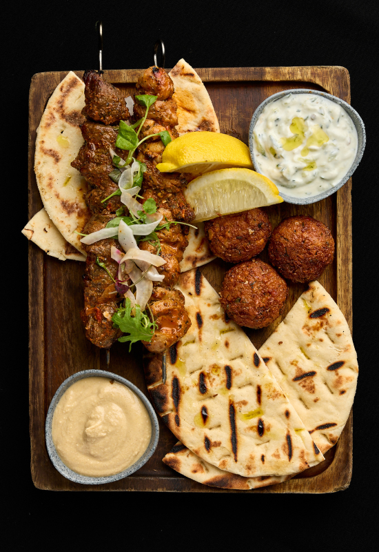A wooden board with grilled pita bread, skewered meat, falafel balls, lemon wedges, sliced onions, fresh herbs, hummus, and tzatziki sauce—an inviting spread inspired by Night Feast 2025 at Brisbane Powerhouse.