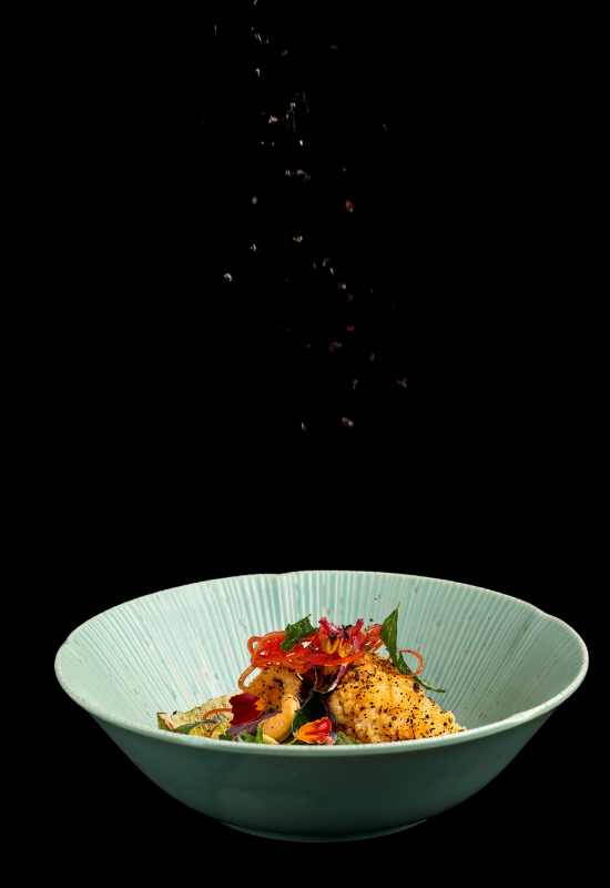 A gourmet dish in a light blue bowl sits against a black background at Night Feast 2025 Brisbane Powerhouse, garnished with herbs and thinly sliced vegetables, as seasonings are sprinkled from above.