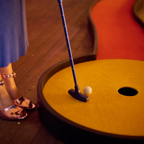 A person in metallic pink heels and a blue skirt stands on a mini-golf course at Brisbane Powerhouse, putting a golf ball toward a hole on a yellow surface with a red path, evoking the playful style of Kaylene Whiskey.