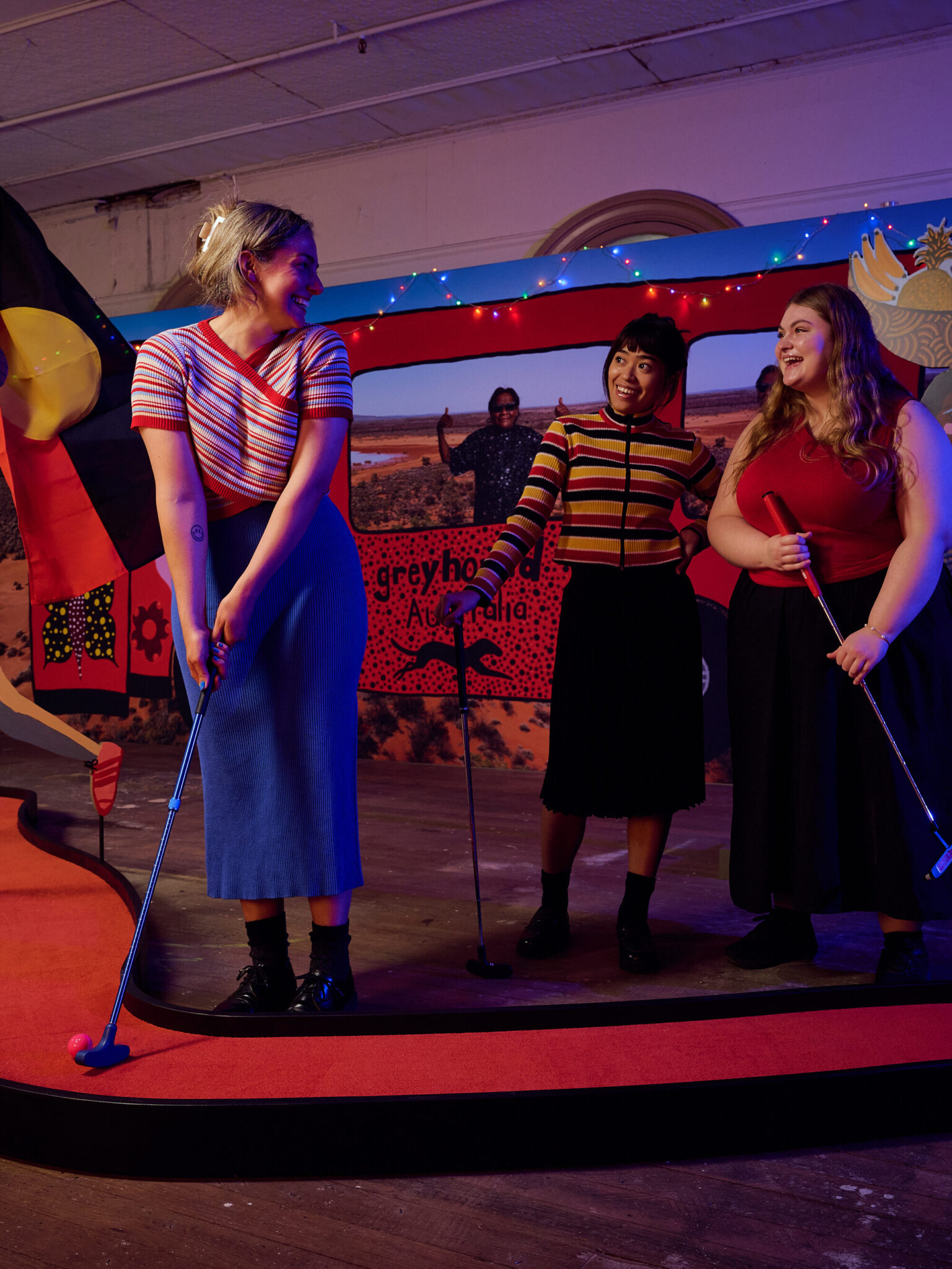 Three women play mini-golf indoors on a colorful, winding course with cartoon-style decorations, including a mural inspired by Kaylene Whiskey at Swingers inside Brisbane Powerhouse. Warm lighting and string lights create a playful, festive atmosphere.