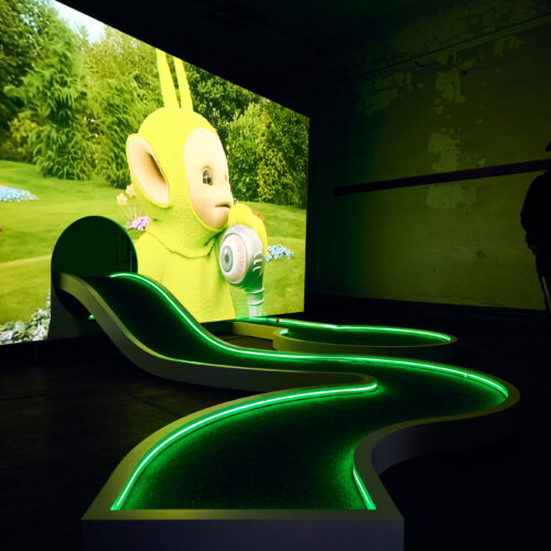 A wavy mini-golf course with green LED lights stands in front of a large screen showing a Teletubby character. Reminiscent of a Miranda July installation, the scene glows in the dimly lit Brisbane Powerhouse space.