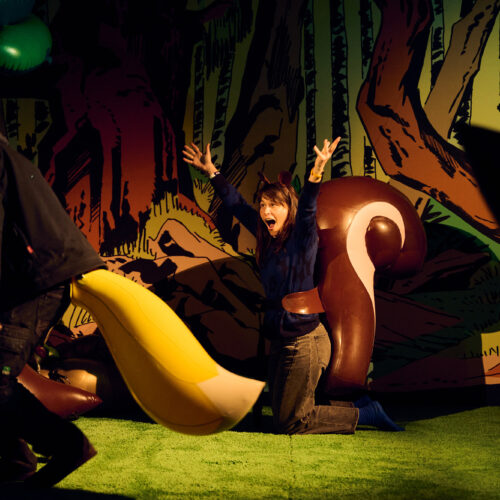 A person kneels on fake grass with arms raised, wearing squirrel ears and posing excitedly near a large, cartoonish squirrel tail prop—an offbeat scene worthy of Miranda July. Another person with a yellow tail costume passes by a Brisbane Powerhouse forest backdrop.