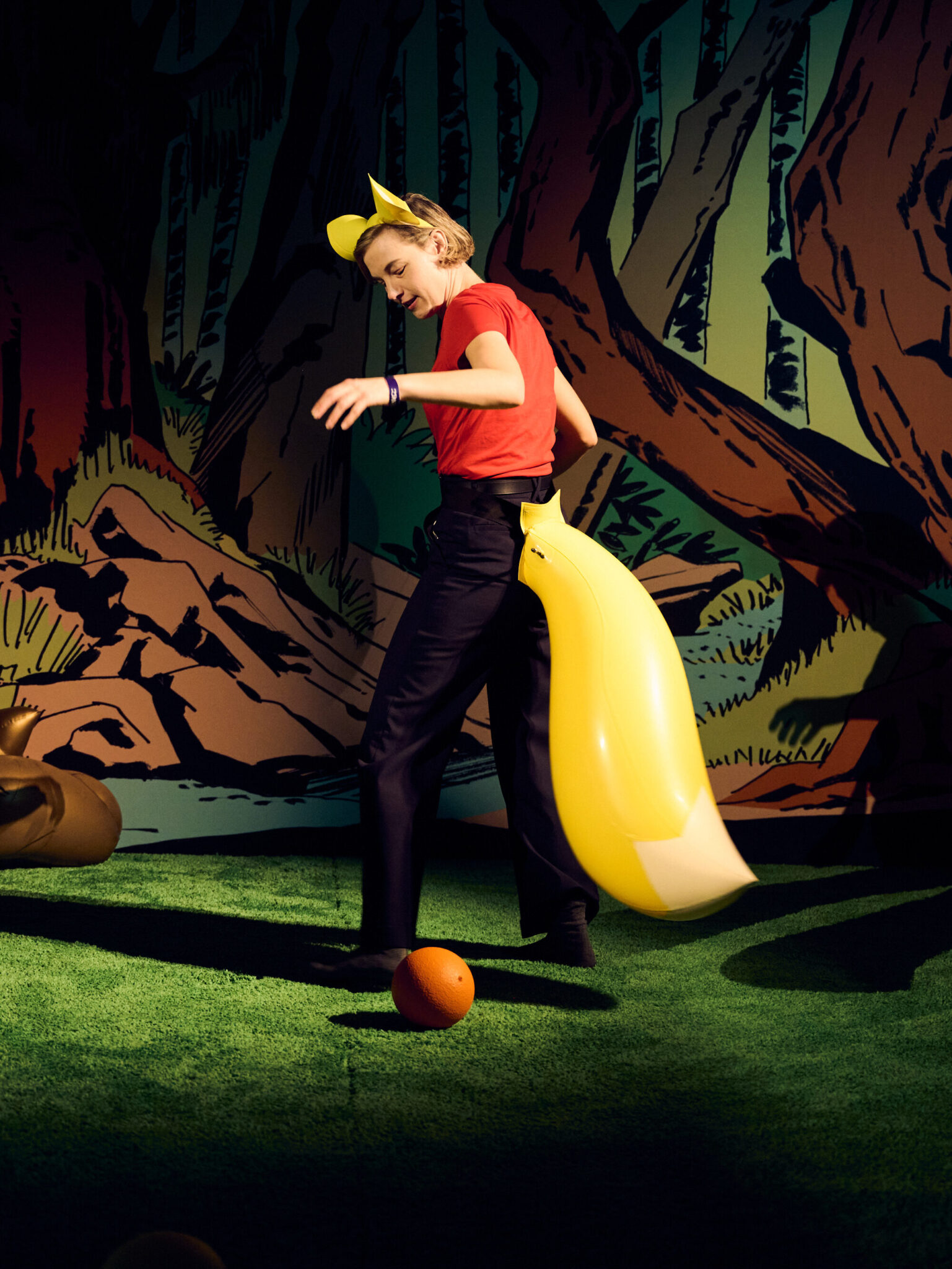 A person dressed in black pants, a red shirt, yellow fox ears, and a yellow fox tail stands on artificial grass at Saeborg Swingers Brisbane Powerhouse, with two orange balls on the ground and a cartoon forest backdrop.