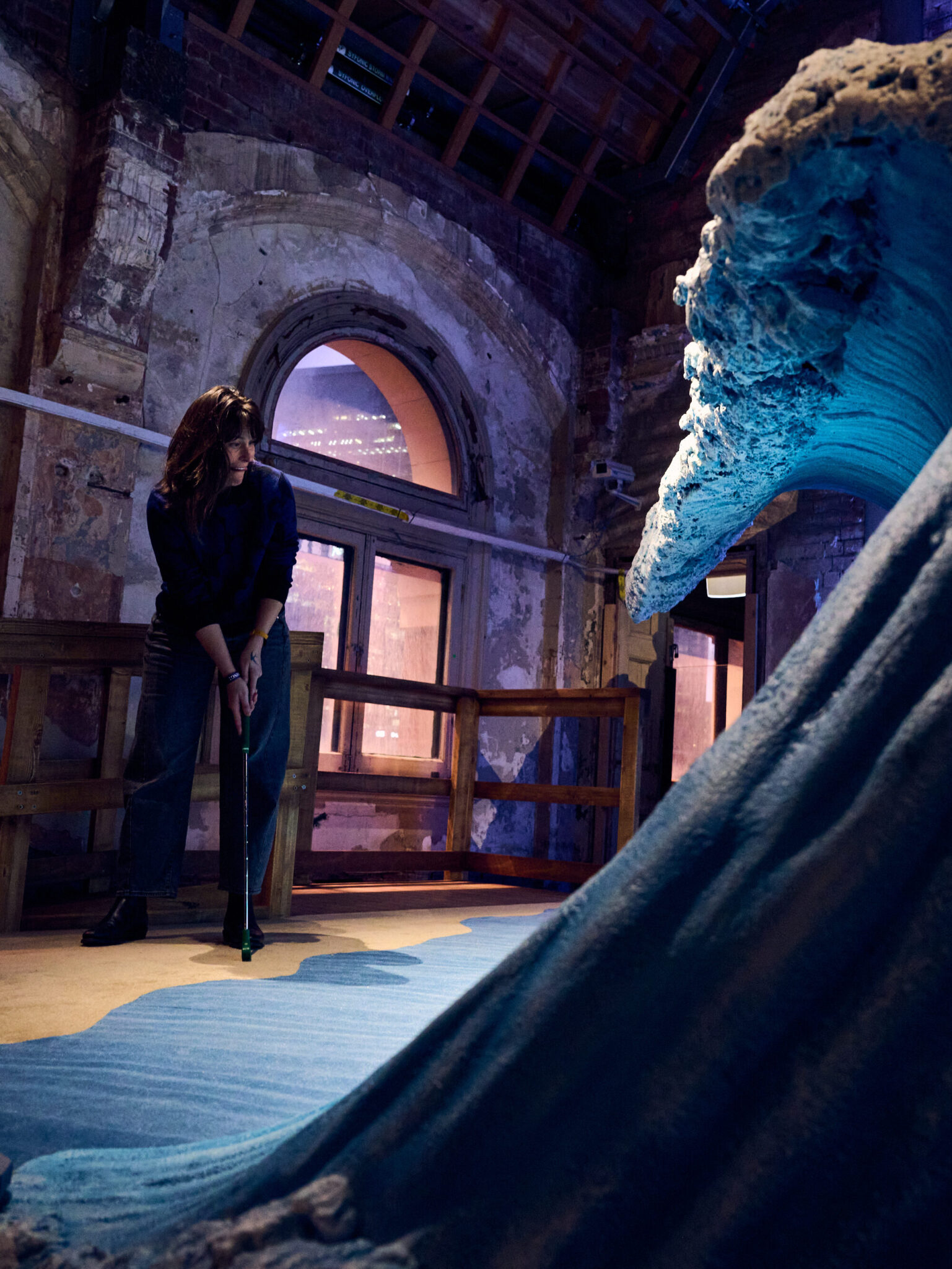 A person plays mini-golf indoors next to a large, detailed wave sculpture at Brisbane Powerhouse, inside a rustic building with arched windows and exposed brick walls.
