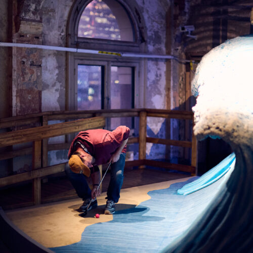 A person in a red jacket and yellow cap bends down to touch a large, realistic blue wave sculpture indoors at Brisbane Powerhouse, surrounded by rustic, weathered walls and windows.