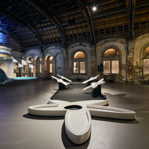 A large, open indoor space with high arched windows, exposed brick walls, and a mini-golf course featuring sculptural white obstacles. A whale and a “Wave of Fortune” sign are visible at Swingers Brisbane Powerhouse.