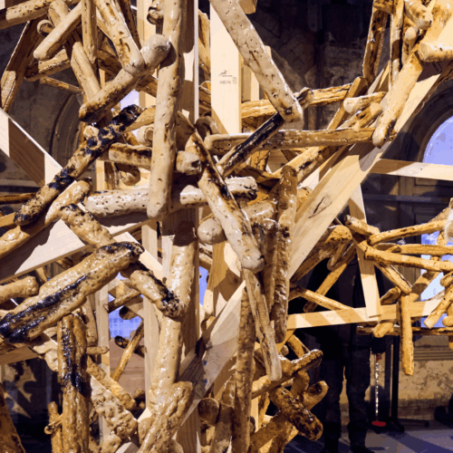 A person with light hair examines an intricate wooden art installation by Nabilah Nordin, reminiscent of her "Swingers" exhibit at Brisbane Powerhouse, made of interlocking beams and branches in a space with arched windows and rustic walls.