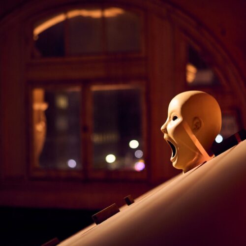 A mannequin head with an open mouth is positioned on a slanted surface at night, with a large, arched window and blurred city lights in the background. The scene is dimly lit with a warm, reddish glow.