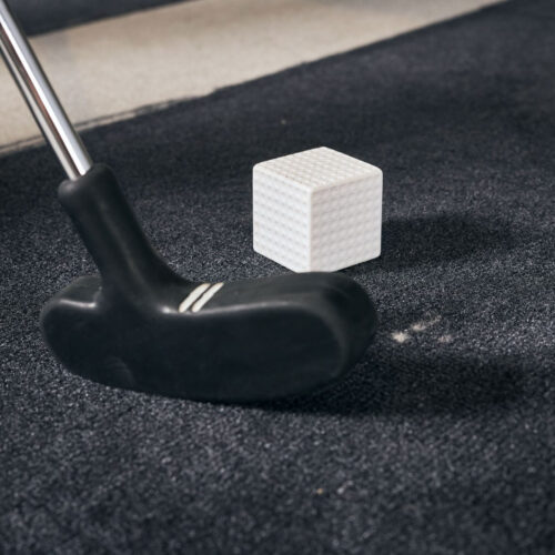 A black golf putter is positioned next to a white dimpled cube, resembling a golf ball in texture, on a dark carpeted surface with beige stripes.