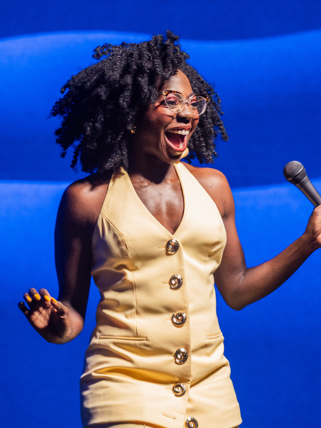 A woman with natural curly hair, wearing glasses and a yellow sleeveless outfit, smiles widely while holding a microphone, standing in front of a blue wavy background.