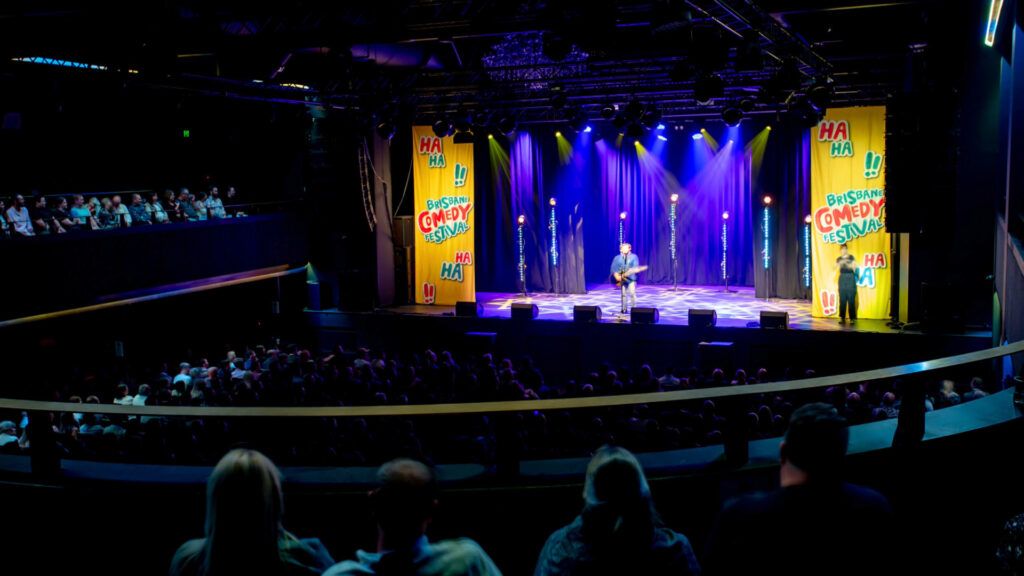 A comedian performs on a brightly lit stage at a comedy festival in front of a large seated audience in a dark theater. Colorful banners with “Britsish Comedy Festival” and “HA HA” decorate the stage.