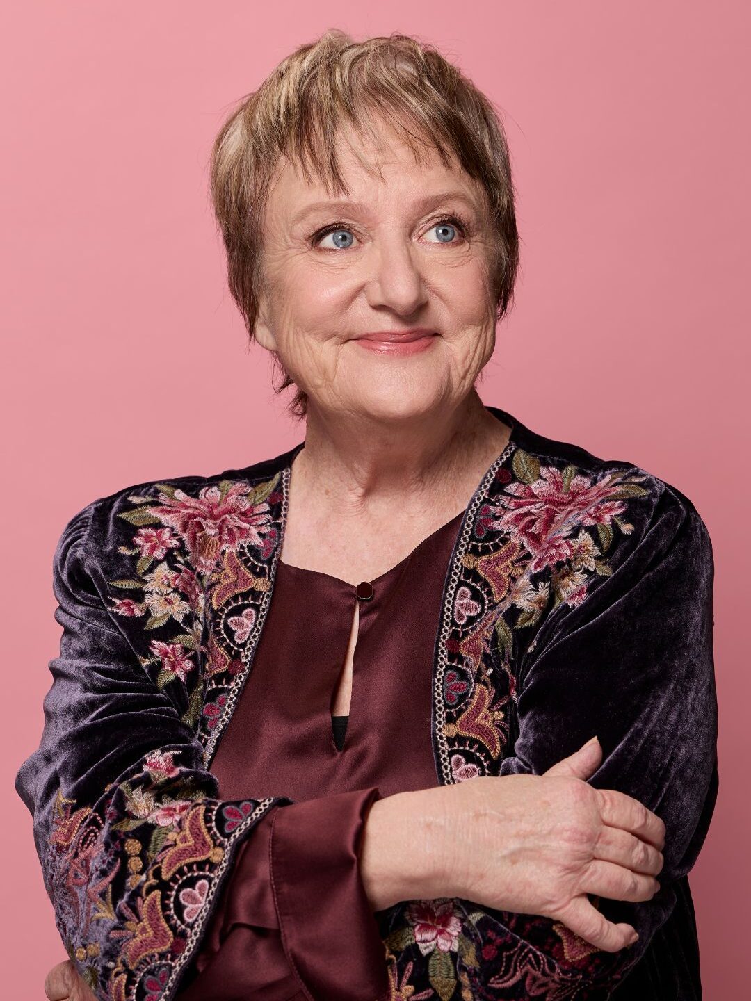 Denise Scott, an older woman with short light brown hair, smiles while looking off-camera. She wears a burgundy blouse and a dark velvet jacket with floral embroidery, standing against a pink background with her arms gently crossed.