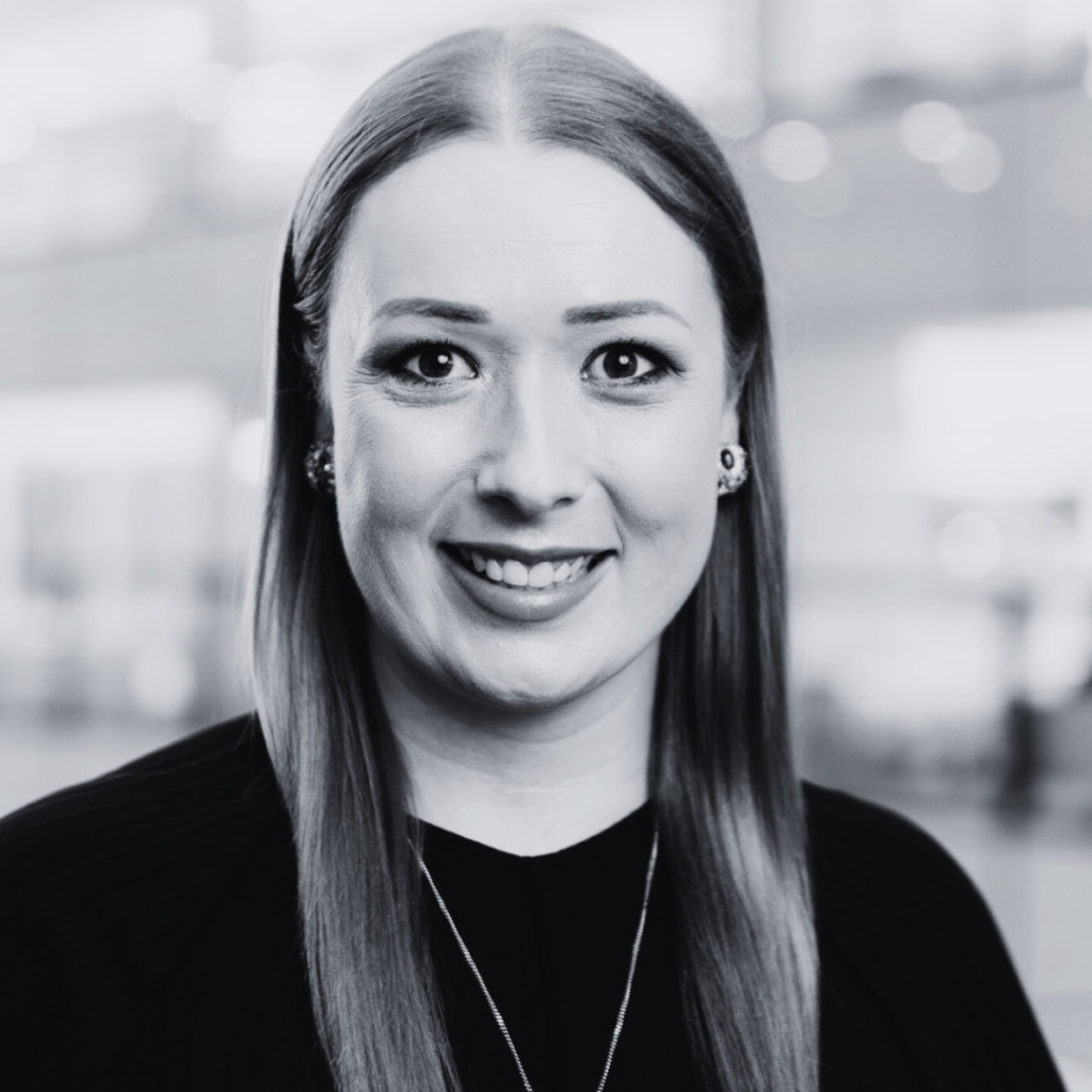 A woman with straight, light-colored hair, wearing a dark top and earrings, smiles at the camera. The blurred background suggests an indoor office setting. This black-and-white image captures a member of Our Team - Brisbane Powerhouse.