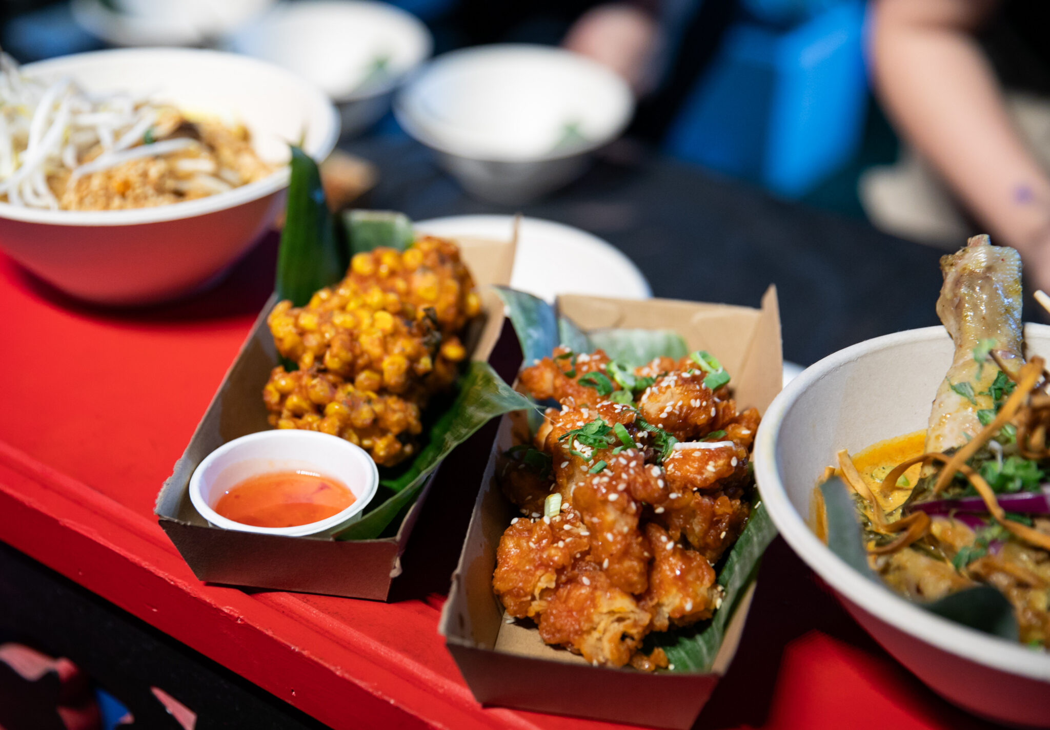 Close-up of various Asian street food dishes, including fried corn fritters with dipping sauce, glazed fried chicken garnished with sesame seeds and herbs, and noodle soup bowls in the background.