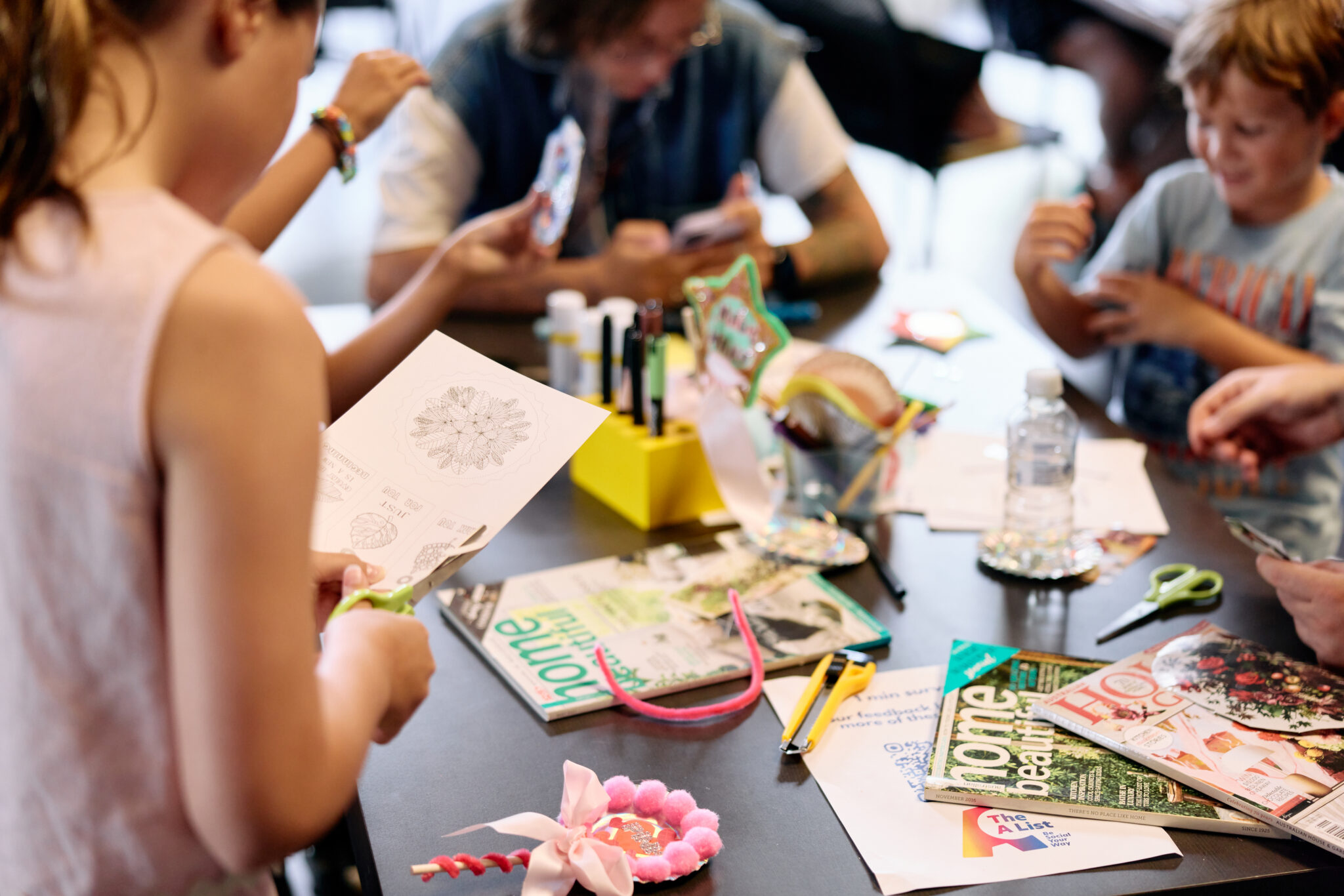 Children and adults sit around a table covered with art supplies, magazines, papers, markers, and craft materials, creating various art projects together in a lively, creative setting.