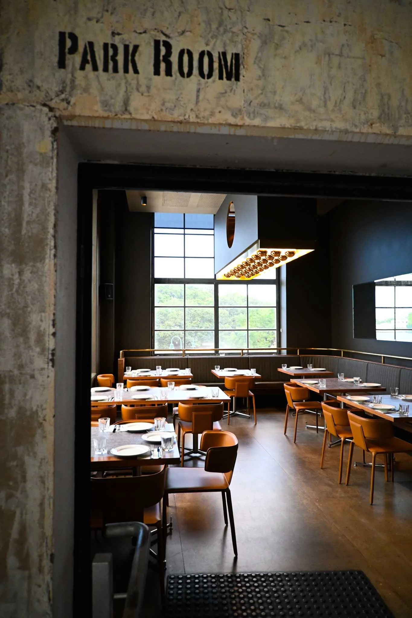 A dining room labeled PARK ROOM with large windows, modern lighting, and neatly set tables with brown chairs, viewed from an open doorway.