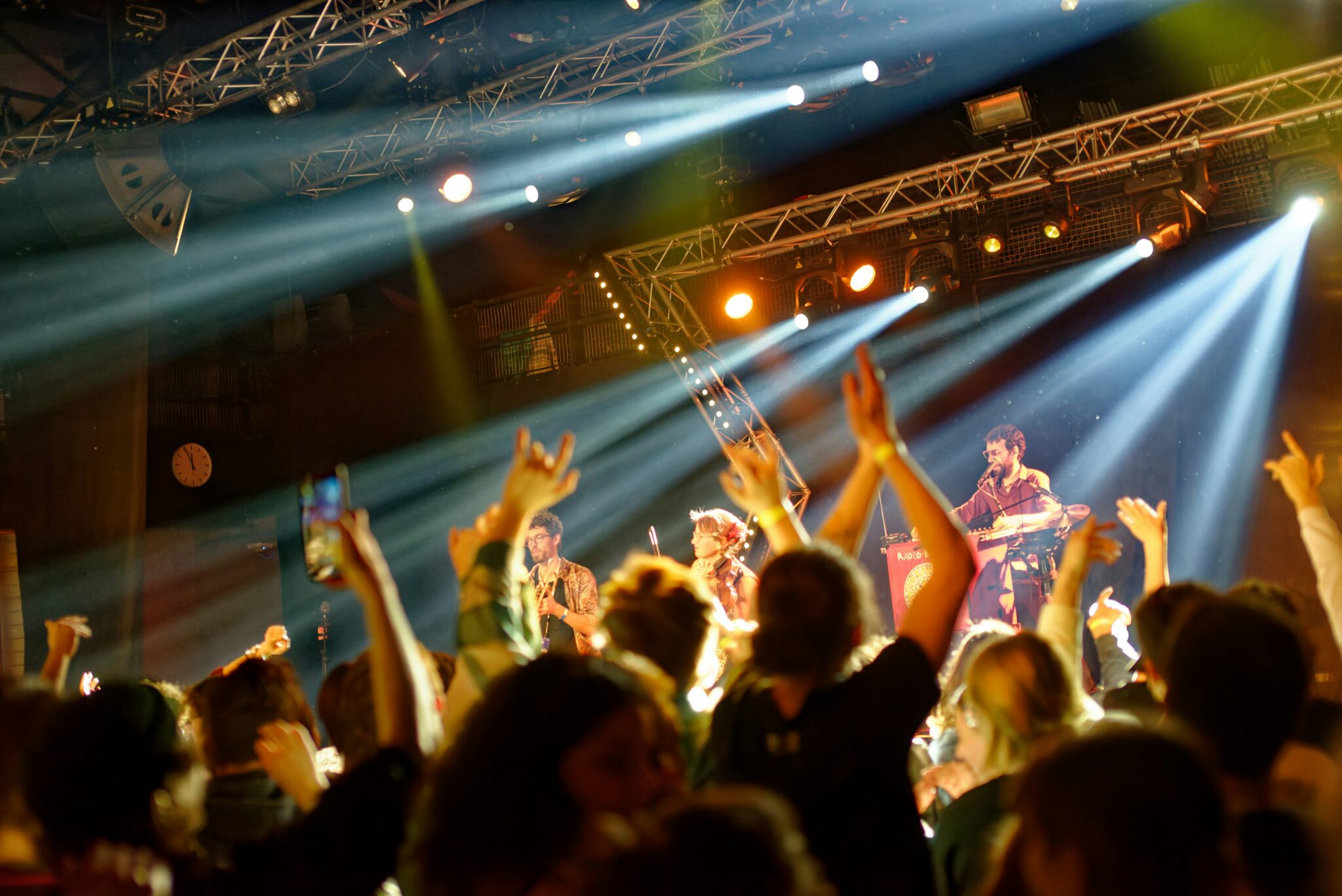 A lively concert scene with a cheering crowd, raised hands, and bright stage lights illuminating musicians performing on stage under a metal truss.