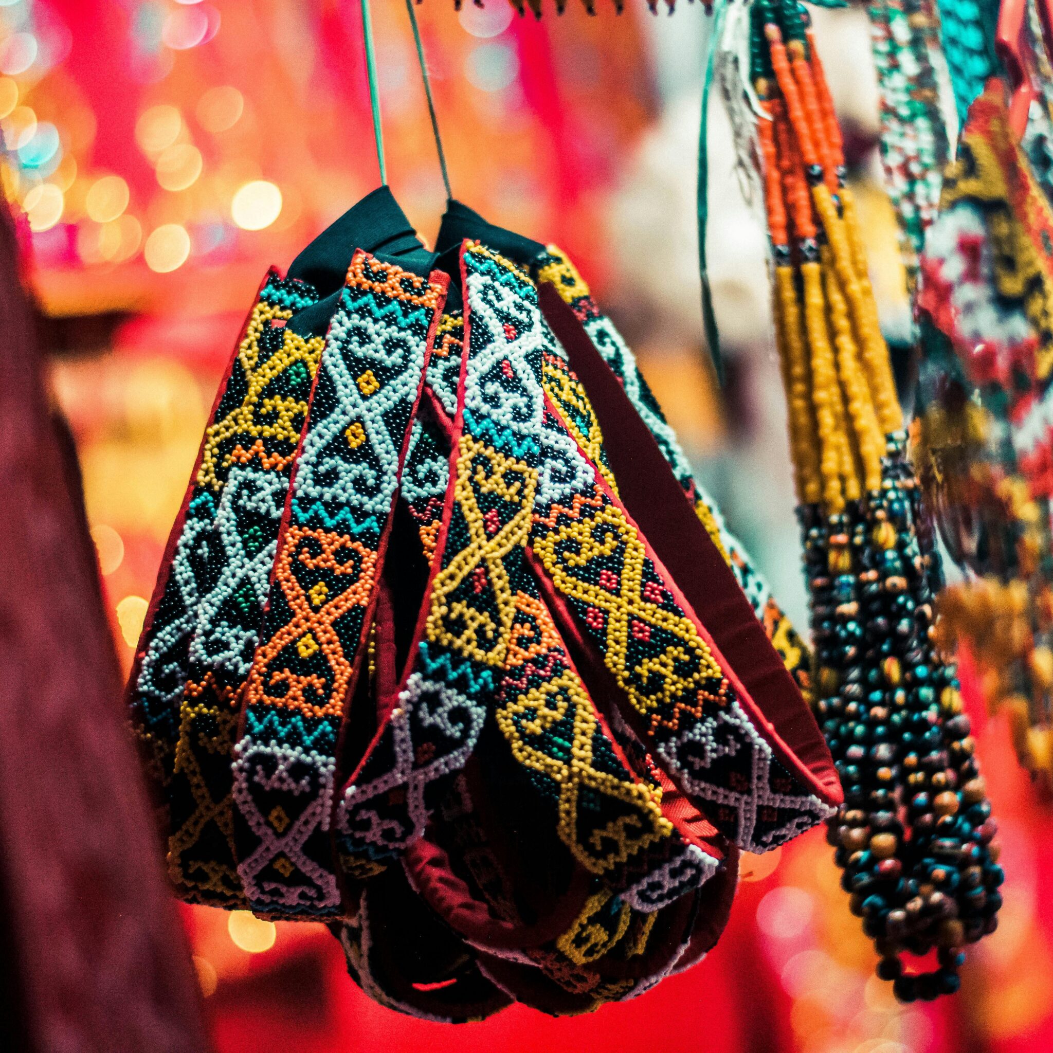 A close-up of intricately woven, colorful beaded bracelets with geometric patterns hanging on display, surrounded by other beaded jewelry against a vibrant, blurred red background.