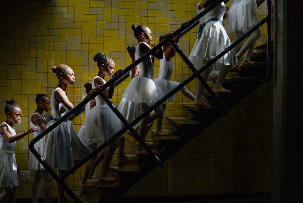 A group of young ballerinas in white tutus and leotards ascend a dimly lit staircase beside a yellow-tiled wall, holding the railing in single file—capturing the quiet beauty featured at the World Press Photo Exhibition 2025 Brisbane Powerhouse.