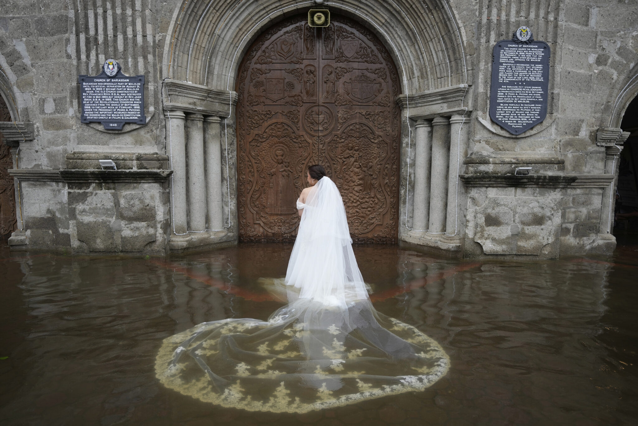 A bride in a white gown and veil stands in knee-deep floodwater before ornate church doors, stone walls and plaques behind her—a striking image from the World Press Photo Exhibition 2025 at Brisbane Powerhouse.