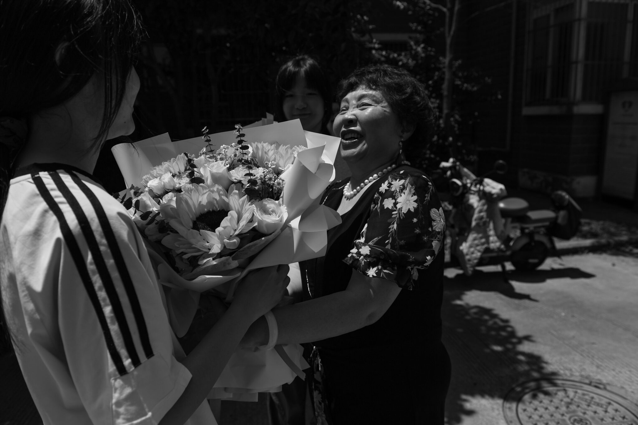 An elderly woman smiles joyfully as she receives a large bouquet of flowers from a younger person outdoors, with another standing behind them on a sunny day—capturing moments like those seen at the World Press Photo Exhibition 2025 Brisbane Powerhouse.