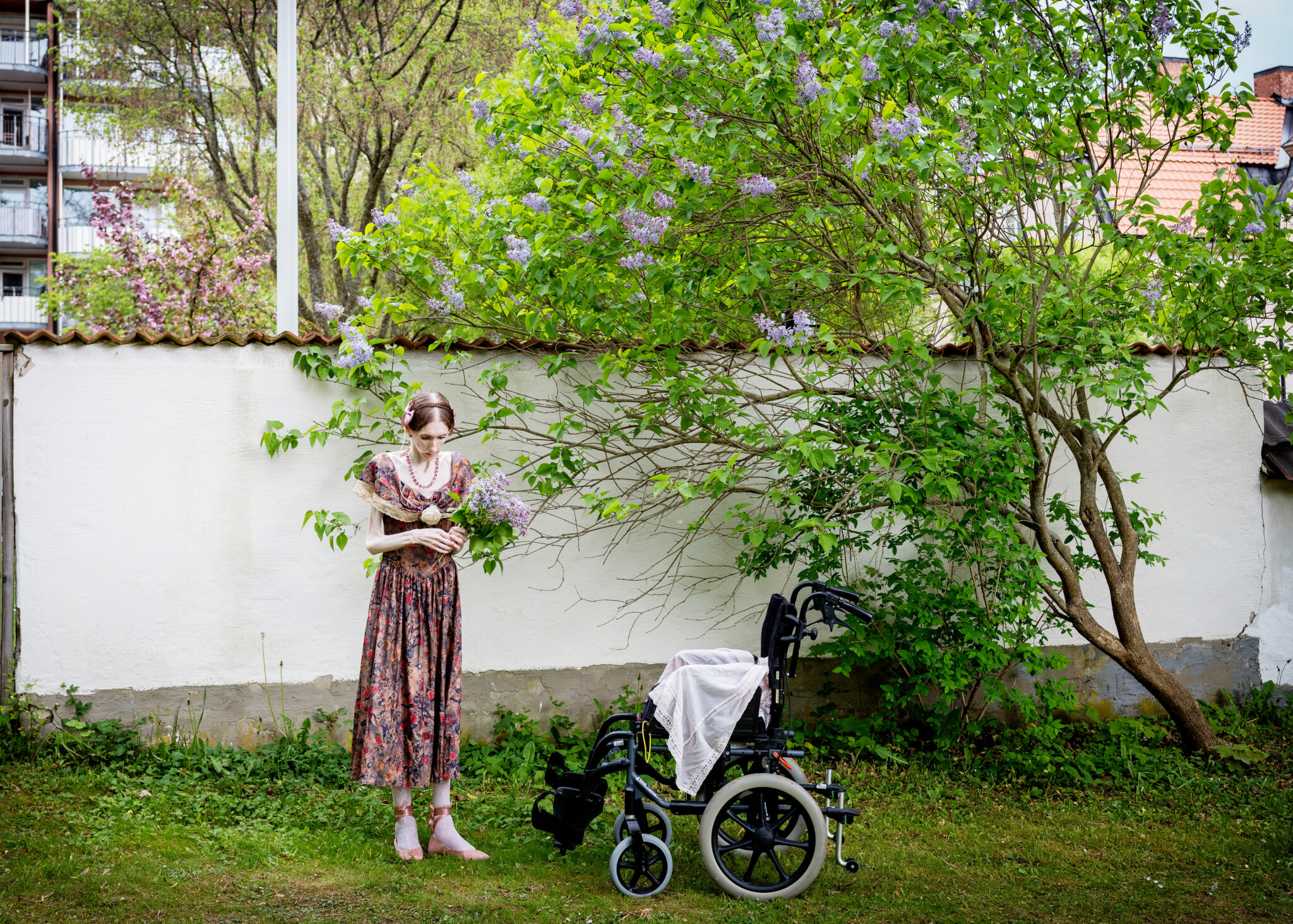 A woman in a floral dress stands in a garden holding lilac flowers near a lilac bush, beside an empty wheelchair. This evocative scene echoes the storytelling seen at the World Press Photo Exhibition 2025 Brisbane Powerhouse.
