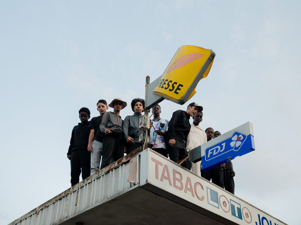A group of young people stand closely together on a rooftop beneath a cloudy sky, with signs reading PRESSE and TABAC LOTO FDJ. Their gathering echoes the anticipation for World Press Photo Exhibition 2025 at Brisbane Powerhouse.