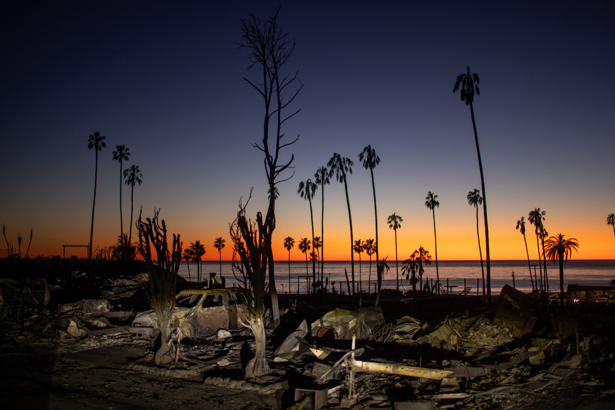 Burnt palm trees, charred debris, and a damaged car stand silhouetted against an orange and blue sunset by the ocean—a striking scene reminiscent of works shown at the World Press Photo Exhibition 2025 Brisbane Powerhouse.