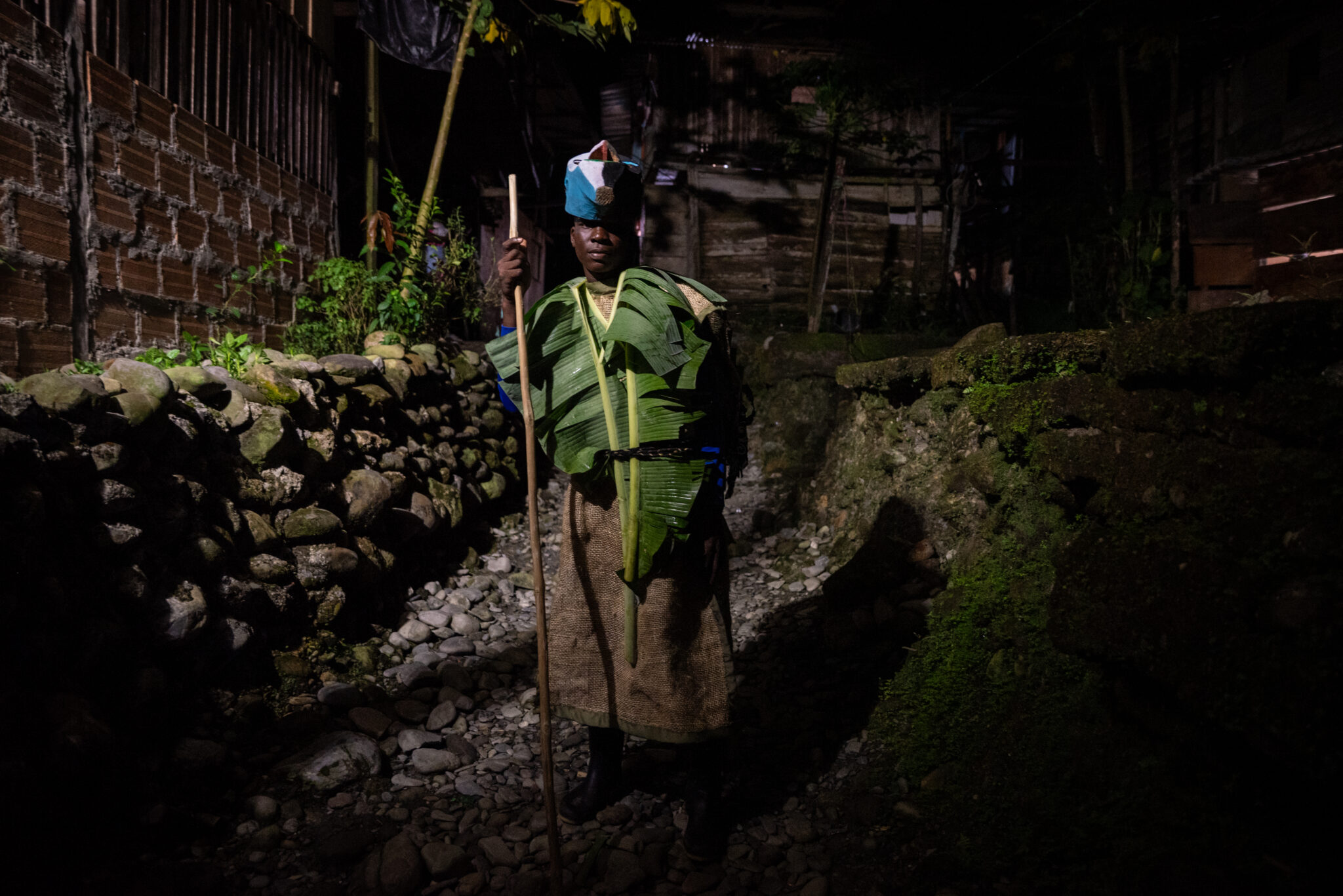 A person stands on a rocky path at night, dressed in clothing made from leaves and natural materials, holding a long staff. Stone walls and wooden buildings surround the scene, their shadows echoing the drama of a World Press Photo Exhibition 2025 Brisbane Powerhouse.