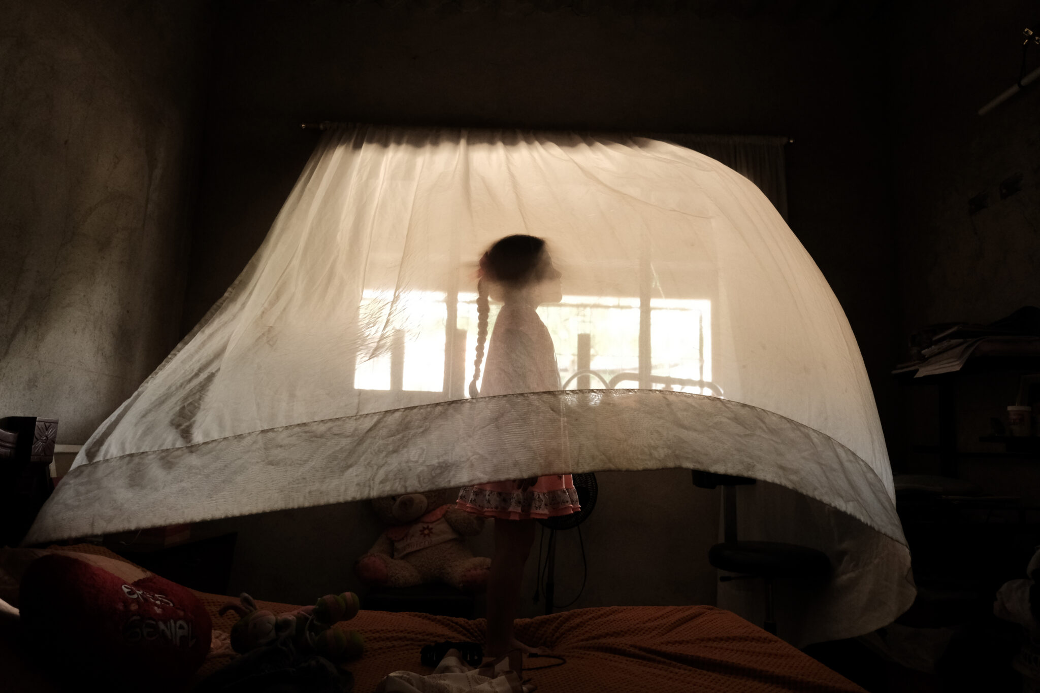 A young child stands on a bed behind a sheer white curtain, softly illuminated by sunlight streaming through a window in a dimly lit room—capturing the quiet moments celebrated at the World Press Photo Exhibition 2025 Brisbane Powerhouse.