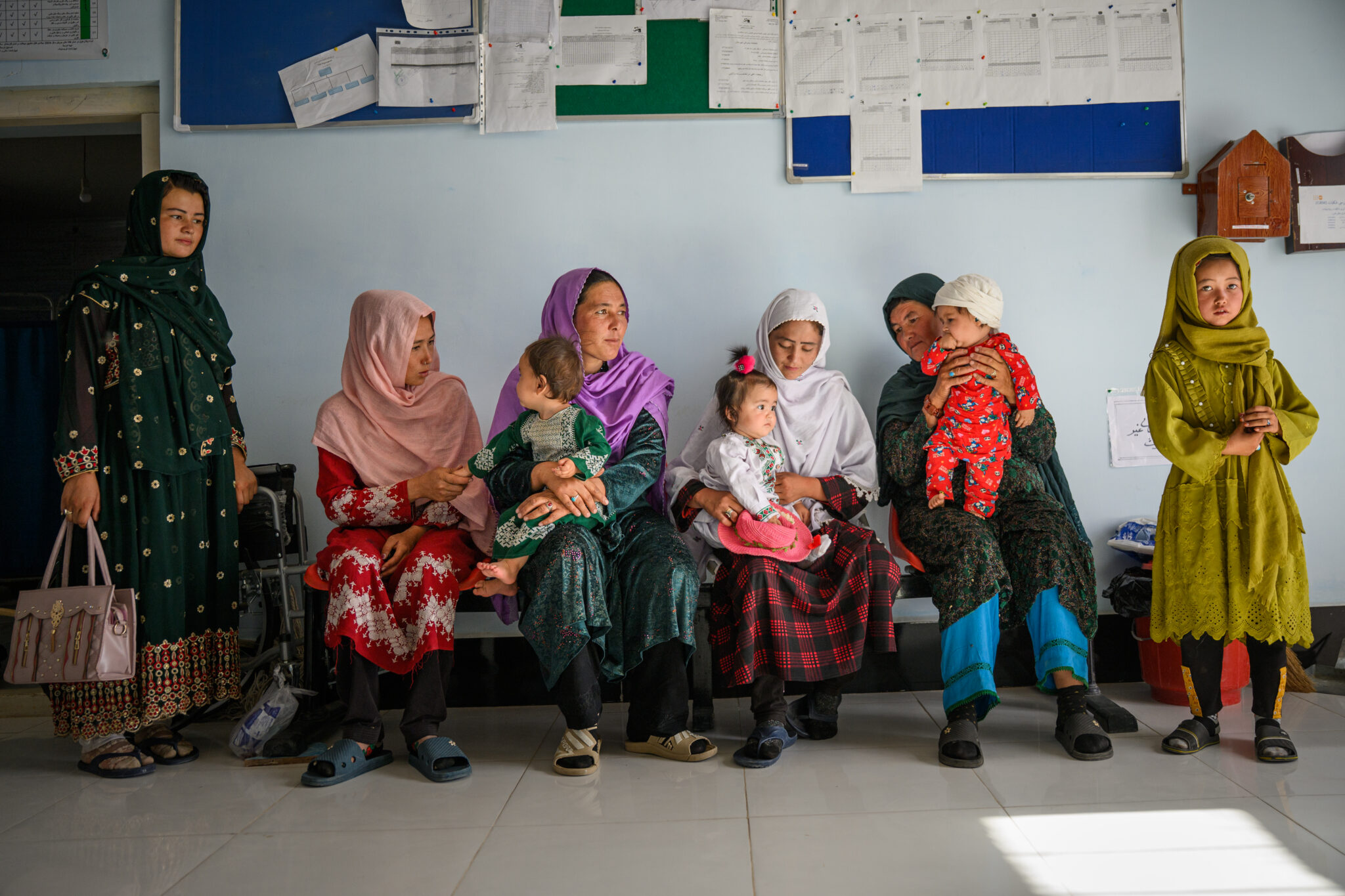 A group of women and young girls, some holding babies, sit or stand in a waiting area with white walls and bulletin boards. Their colorful traditional clothing brightens the scene featured at the World Press Photo Exhibition 2025 Brisbane Powerhouse.
