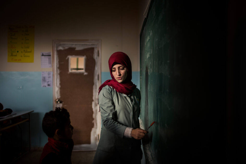 A woman in a red hijab writes on a chalkboard in a dimly lit classroom at the World Press Photo Exhibition 2025 Brisbane Powerhouse, while a young boy stands nearby. Faded walls and educational posters complete the evocative scene.