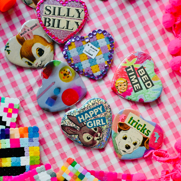 Colorful heart-shaped pins with playful designs and words like SILLY BILLY, BED TIME, HAPPY GIRL, and tricks are arranged on a pink gingham cloth surrounded by bright bead art.
