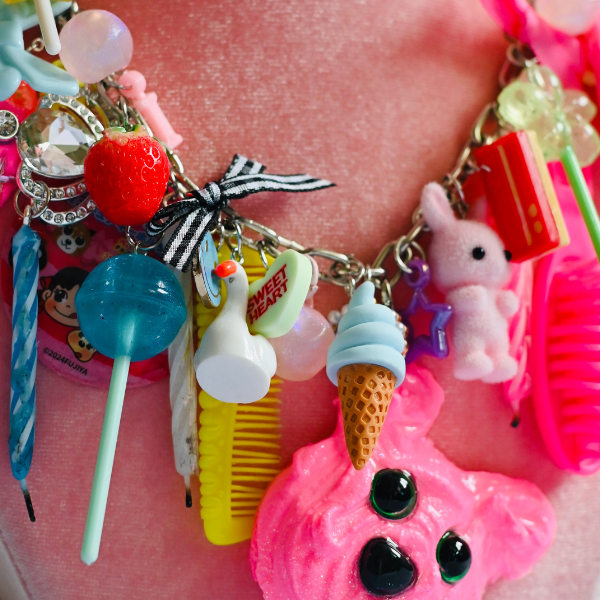 A colorful charm necklace with trinkets including a lollipop, strawberry, ice cream cone, toothbrush, pink bunny, comb, and other cute decorations, displayed on a pink mannequin bust.