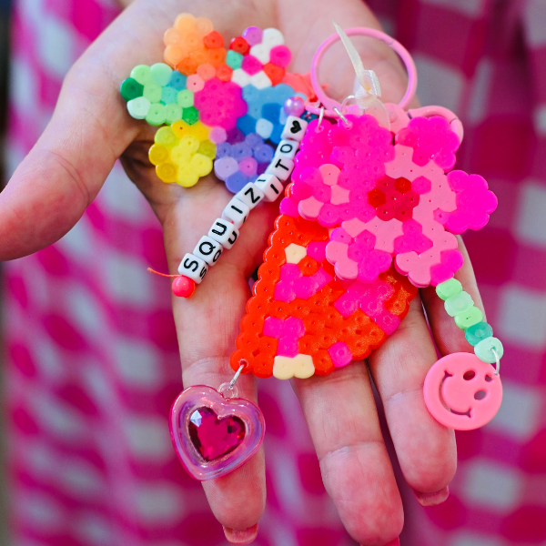 A hand holds colorful pixel bead keychains, including a pink heart, a strawberry, a flower, a smiling face charm, and a beaded string spelling SQUIZITO!, against a pink checkered background.