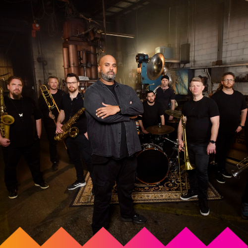 A band of eight men poses in an industrial setting with musical instruments including saxophones, trombones, and drums; the central figure stands with arms crossed, while others stand or sit around him.
