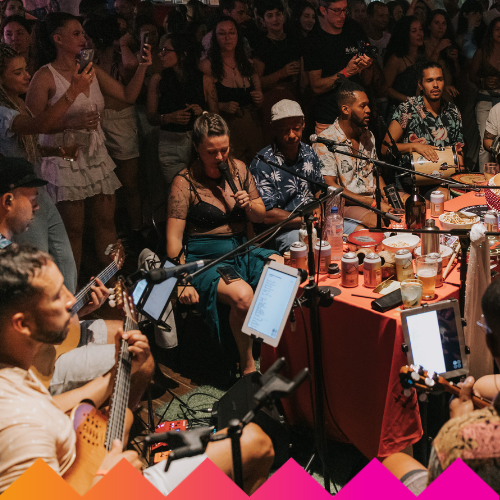 A lively indoor gathering at Ubuntu Social Brisbane Powerhouse with musicians playing guitars and singing around a table, as LED HULA HOOP performers and a festive crowd create an engaging atmosphere.