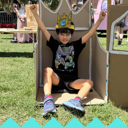 A child wearing a yellow paper crown sits inside a cardboard castle with arms raised and smiling. The child wears a black T-shirt with colorful designs, black shorts, and blue sneakers on green grass at an outdoor event.