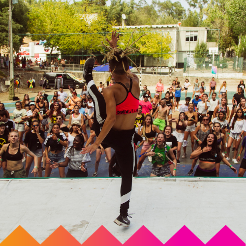 A dance instructor on stage, mid-kick, leads a large, enthusiastic crowd in an outdoor dance class at Ubuntu Social Brisbane Powerhouse. Colorful graphic shapes and an LED hula hoop light up the scene, with trees and buildings in the background.