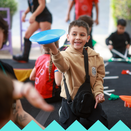 A smiling boy in a tan hoodie and black crossbody bag plays with a blue plastic plate, surrounded by other children at Ubuntu Social Brisbane Powerhouse, where LED hula hoops and laughter light up the lively indoor setting.