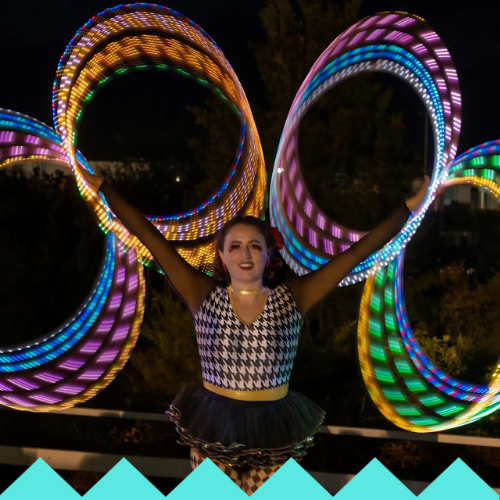 A performer in a black-and-white patterned outfit twirls multiple LED HULA HOOPS, creating colorful light trails at night. She stands smiling outdoors at the Ubuntu Social, Brisbane Powerhouse, with arms raised against a dark background.