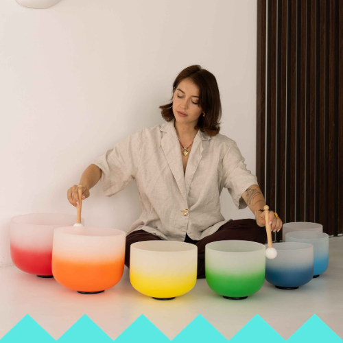 A woman in a beige shirt sits on the floor playing colorful crystal singing bowls with mallets, while an LED HULA HOOP glows nearby, creating a peaceful atmosphere in a modern, minimalist room.