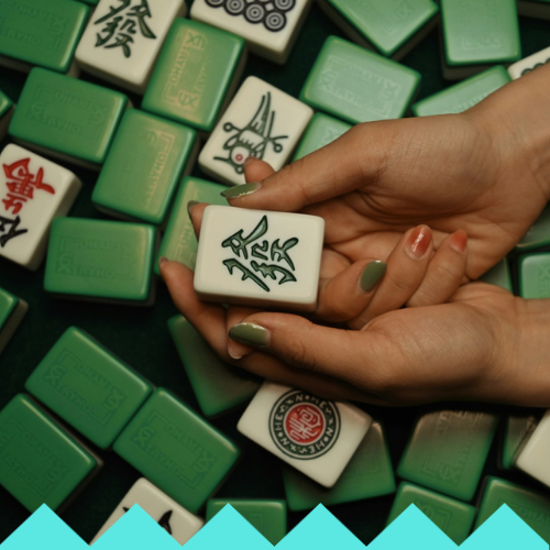 A pair of hands with painted nails holds a green-and-white Mahjong tile above a scattered pile of similar Mahjong tiles on a dark green surface at the LED HULA HOOP - Ubuntu Social Brisbane Powerhouse.