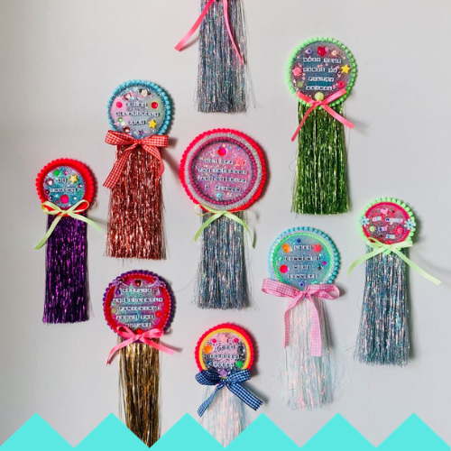 Nine colorful rosette-style decorations with long, shiny tassels and bows are displayed on a white wall. Inspired by the vibrancy of an LED HULA HOOP, each rosette has decorative text in the center and is surrounded by small embellishments.