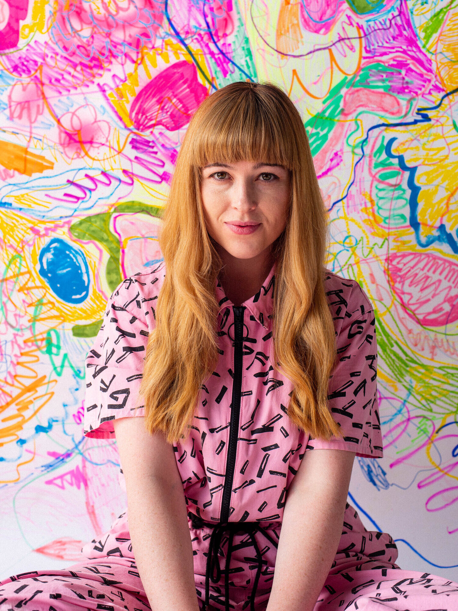 A woman with long blonde hair and bangs, wearing a pink patterned outfit, sits in front of a colorful, abstract, hand-drawn background filled with vibrant swirls, lines, and shapes.