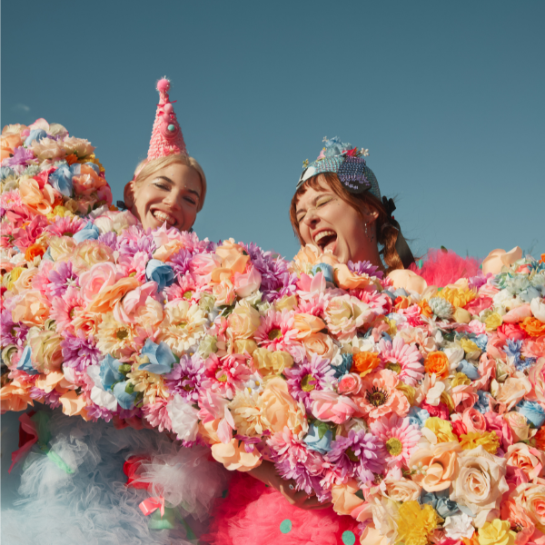 Two people wearing party hats and colorful costumes smile and laugh, surrounded by a large, vibrant arrangement of multicolored artificial flowers against a clear blue sky.