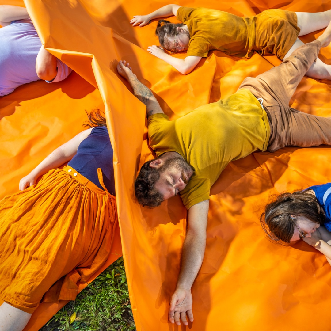Four people lie on an orange sheet spread on grass, dressed in blue, yellow, and orange. Sunlight casts shadows across the scene during Bleachers at Brisbane Festival 2026, creating a relaxed festival atmosphere.